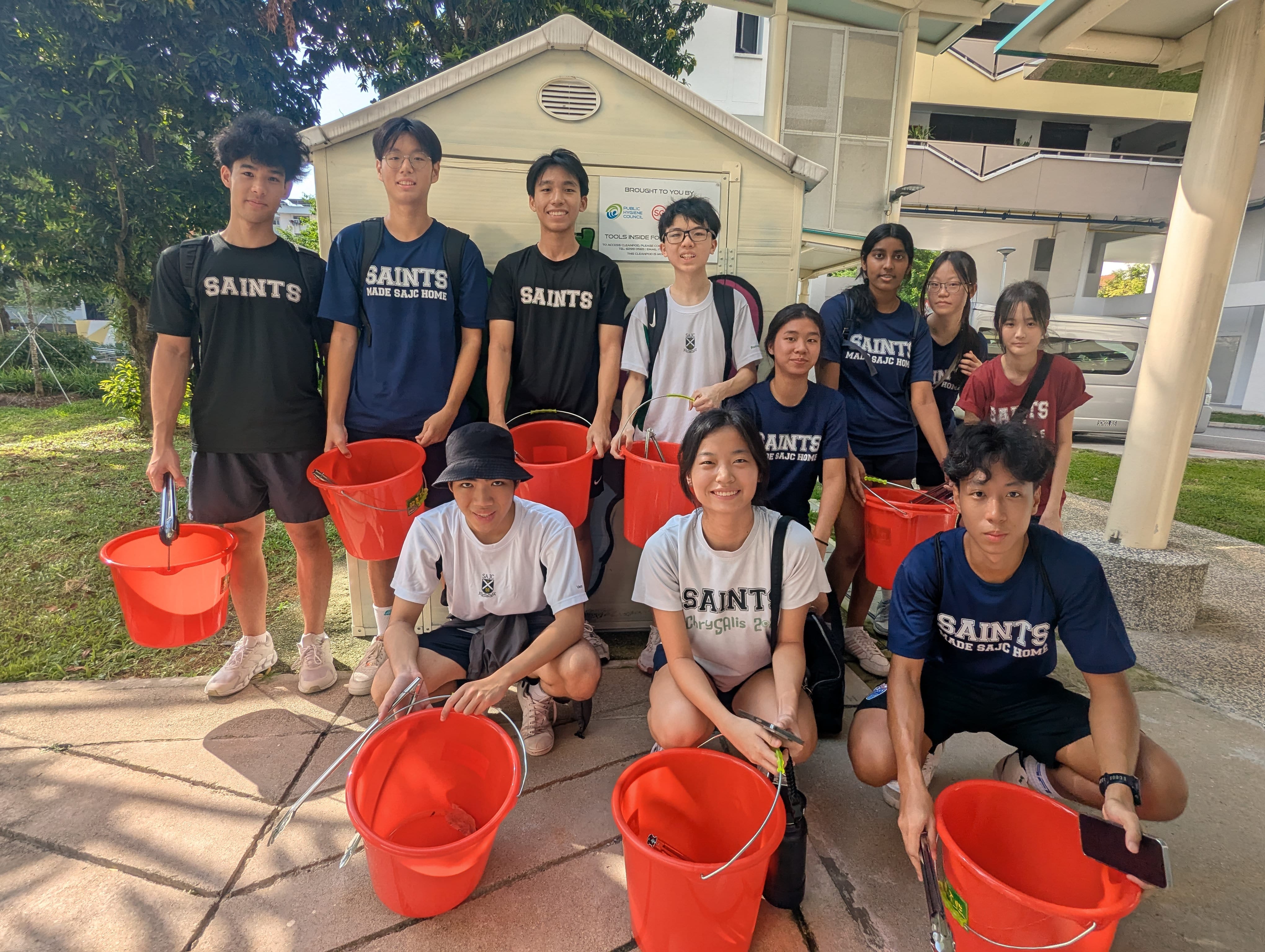 A group of student volunteers posing outdoors holding red buckets and metal trash tongs during a community cleanup activity.